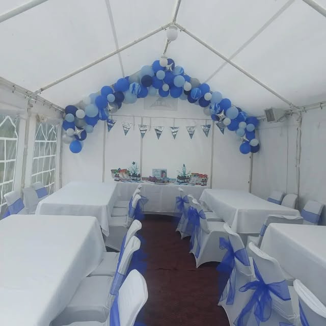 Marquee interior arranged with white-clothed tables, blue chair sashes, and a blue balloon arch.